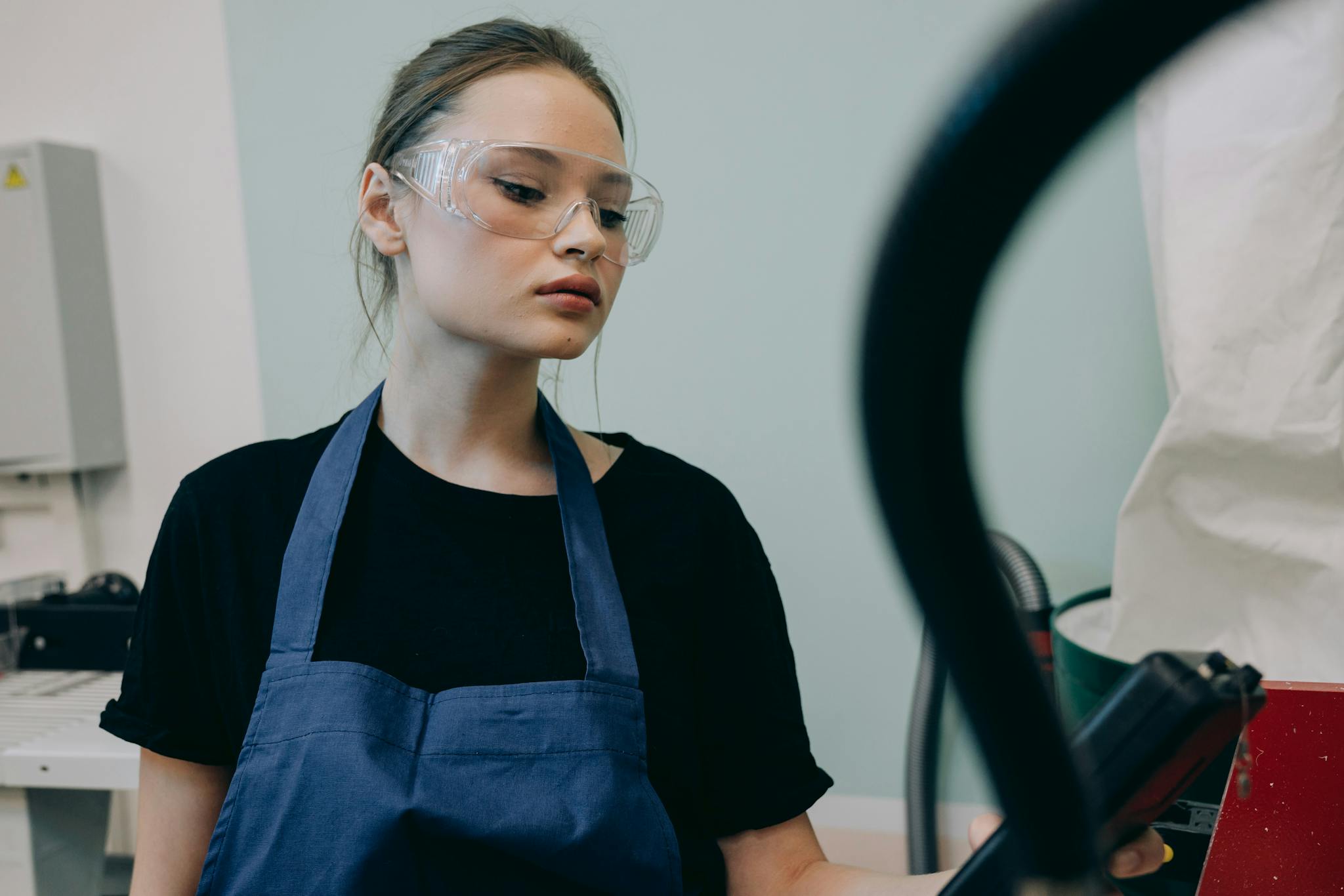 Young woman in a workshop setting, wearing protective goggles and a blue apron, standing indoors.